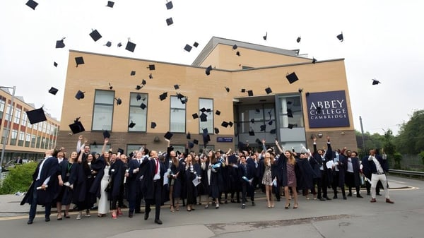 Un grupo de graduados del Abbey College Cambridge lanza sus birretes al aire frente a un edificio de ladrillo.