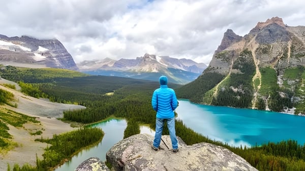 Una persona con un atuendo azul está de pie sobre una roca con vista al paisaje montañoso y al lago cerca de la Abbotsford Senior Secondary School.
