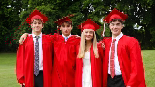 Cuatro estudiantes de la Academy of the New Church llevan togas y birretes rojos y están juntos en un prado con árboles al fondo.