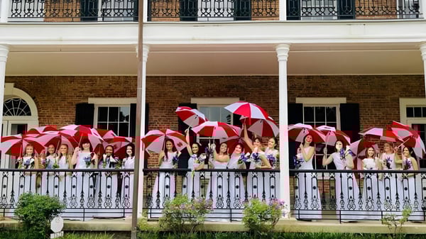 Un grupo de estudiantes con paraguas de colores está en el balcón de un edificio de ladrillo de la Academy of the Sacred Heart.