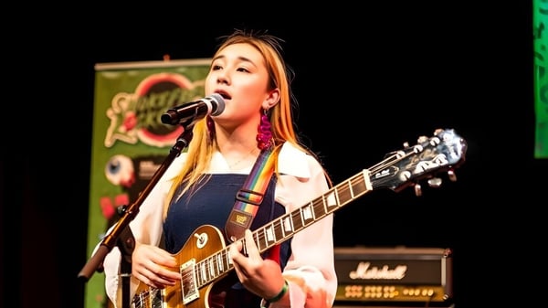 Una estudiante de ACG Sunderland canta y toca la guitarra en una actuación frente a un fondo colorido.