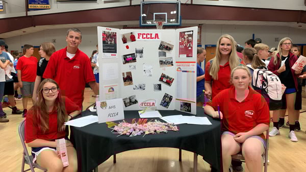 Un grupo de estudiantes de la Acorn High School está en el gimnasio alrededor de una mesa de presentación con carteles y materiales.