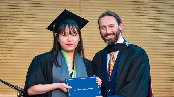 Una graduada en toga de graduación está con un hombre en traje académico y sostiene un diploma en el campus de la ACS International School Cobham.