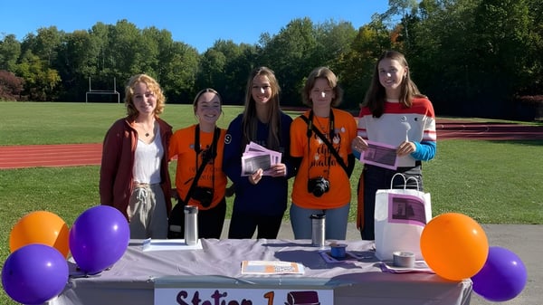 Un grupo de alumnas de la Acton High School está en un campo de césped frente a árboles entre globos de colores y decoraciones.