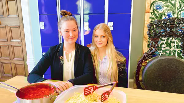 Dos estudiantes de la Adcote School están sentadas juntas en una mesa y comen una comida en una habitación con pared azul y plantas.