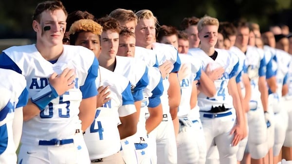 Un grupo de jóvenes atletas está en fila con uniformes blancos y azules en el campo deportivo de Adrian High School.