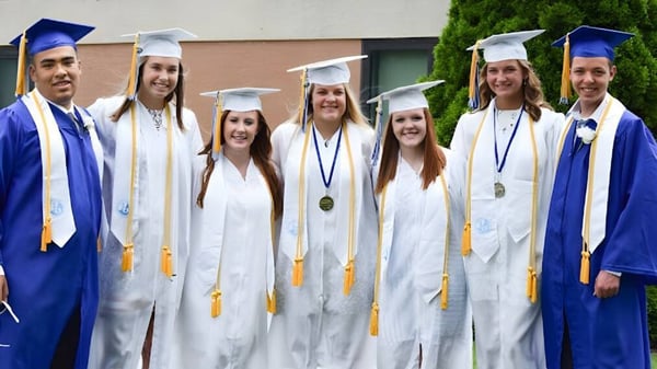 Un grupo de graduados de Adrian High School está en túnicas de graduación azules y blancas frente a un edificio celebrando su graduación.