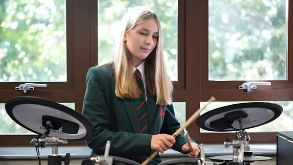 Una estudiante de la Akeley Wood School toca la batería en un interior acogedor con una ventana de fondo.