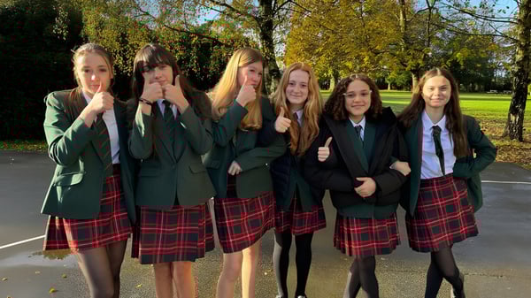Un grupo de estudiantes de la Akeley Wood School está en uniforme escolar juntos en un parque con árboles y plantas.