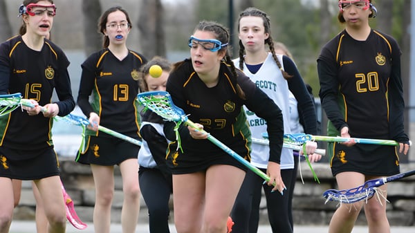 Un grupo de estudiantes del Albert College está con palos de lacrosse en un campo con árboles al fondo.
