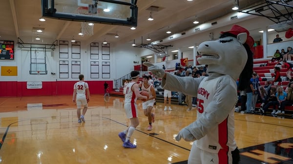 Estudiantes de la Albuquerque Academy juegan baloncesto en el patio escolar con la mascota y espectadores en las gradas.