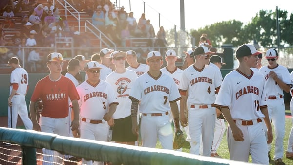 Un grupo de jugadores de béisbol de la Albuquerque Academy está de pie en el campo con espectadores al fondo.