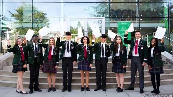 Estudiantes en uniformes escolares están de pie frente a un moderno edificio de vidrio en el campus de la Albyn School sosteniendo papeles o carteles.