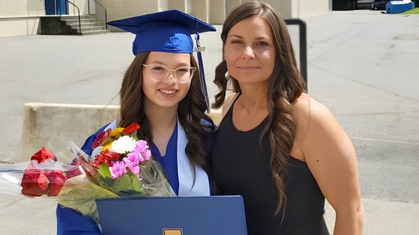 Una estudiante de la Aldergrove Community Secondary School está en una toga de graduación azul junto a una mujer mayor con flores frente al edificio escolar.