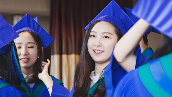 Dos alumnas de la Alexander Academy celebran en togas de graduación azules frente a una cortina roja su graduación.