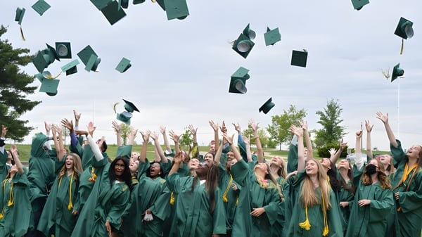 Los graduados de la Alexandra Hills State High School lanzan sus birretes al aire frente a árboles y un cielo nublado.