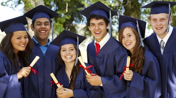 Graduadas y graduados de la All Saints Catholic Secondary School están en sus togas frente a árboles.