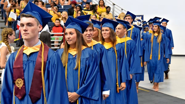 Un grupo de graduados de la All Saints High School en togas azules y amarillas está junto a una multitud.