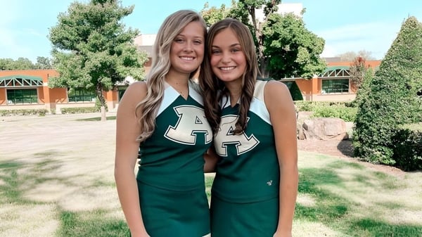 Dos estudiantes de la Alma High School están en camisetas verdes en un campo con árboles y edificios al fondo.