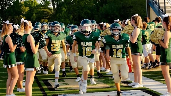 Un grupo de futbolistas en camisetas verdes y blancas están en el campo deportivo de la Alma High School.