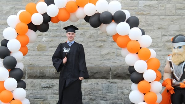 Un estudiante de la Almonte & District High School está vestido con una toga de graduación bajo un arco de globos coloridos frente a una pared de ladrillos.
