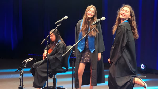 Tres estudiantes de la Alpha School están en ropa de graduación en el escenario y hablan en micrófonos frente a un fondo iluminado en azul.