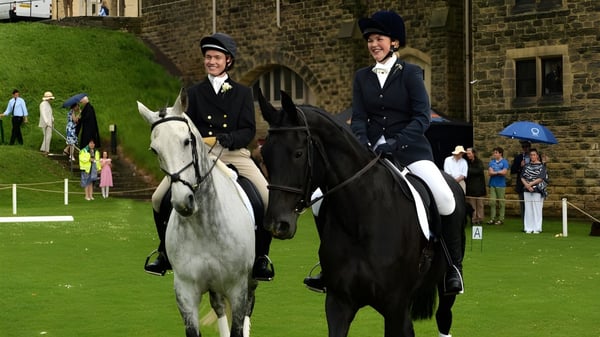 Dos jinetes en ropa formal están con caballos en una pradera frente a un edificio de ladrillo del Ampleforth College.