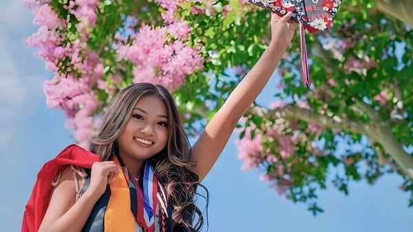 Una joven con cabello largo y oscuro levanta los brazos sonriendo rodeada de flores rosas en el terreno del Anaheim Union High School District.