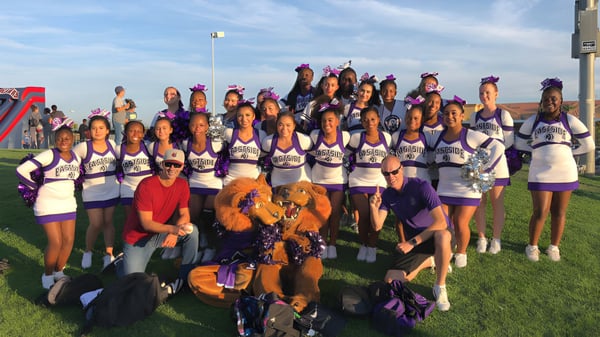 Un grupo de porristas en uniformes morados y blancos está en el campo deportivo del Antelope Valley Union High School District frente a un cielo azul.