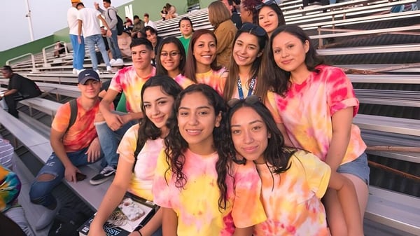 Un grupo de estudiantes riendo en camisetas de Tie-Dye coloridas se sienta en las gradas del campus del Antelope Valley Union High School District con vista al campo deportivo.