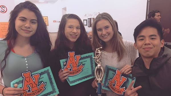 Un grupo de cuatro estudiantes de la Apple Valley Christian School sostiene carteles coloridos en un interior.