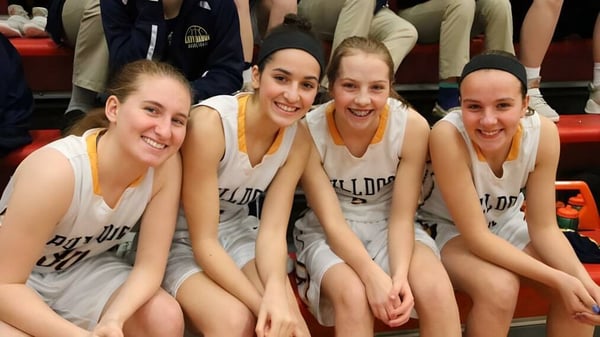 Cuatro estudiantes de Aquin High School están sentadas juntas como equipo de baloncesto y sonríen.