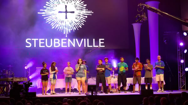 Estudiantes de Aquin High School están en el escenario frente a una gran instalación de luz en forma de cruz con la inscripción STEUBENVILLE.