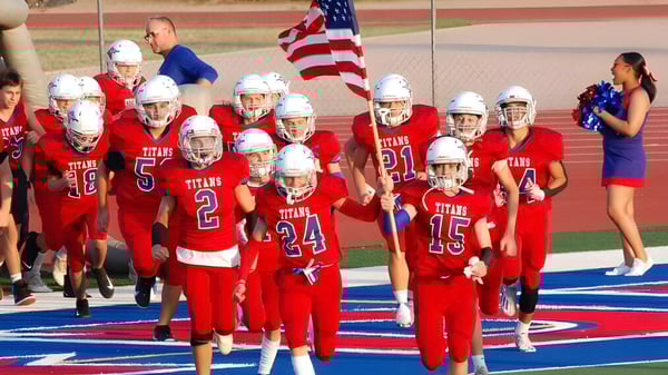 Un grupo de jóvenes jugadores de fútbol americano en camisetas rojas llevan la bandera estadounidense al entrar al campo en el terreno de la Arcadia High School.