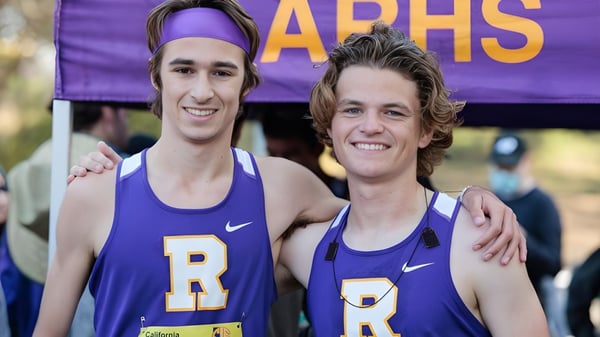 Dos estudiantes de la Archbishop Riordan High School están frente a un banner lila en uniformes deportivos.