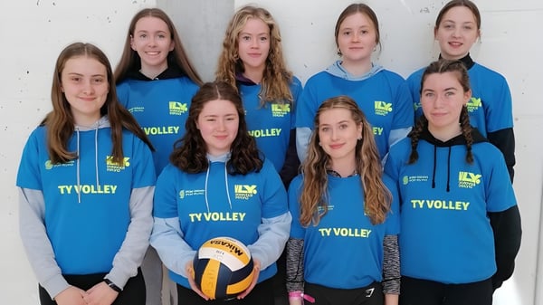 Un grupo de jóvenes jugadoras de voleibol en camisetas azules posan frente a una pared blanca en el campus de la Ardscoil Mhuire.