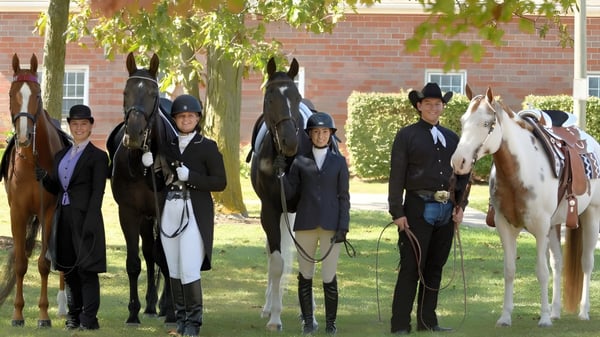 Estudiantes de la Ardscoil Mhuire están en ropa de montar formal junto a caballos en el prado frente a un edificio de ladrillo.