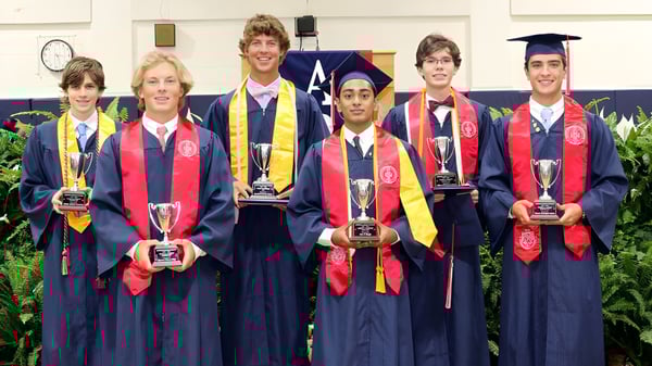 Los graduados de la Arendell Parrott Academy en vestimenta académica sostienen trofeos durante la ceremonia de graduación frente a un fondo verde.