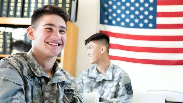 Dos militares sonrientes en uniforme están frente a la bandera americana en el terreno de la Army and Navy Academy.