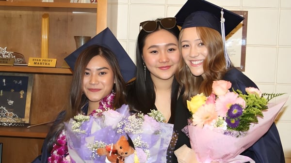 Tres egresadas de la Arroyo Pacific Academy en togas sosteniendo ramos de flores en un entorno hogareño.