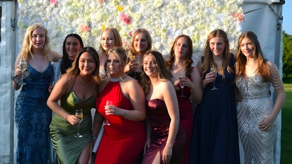 Un grupo de jóvenes mujeres en vestidos de gala se encuentra frente a una colorida pared de flores en el campus de la Ashford School.
