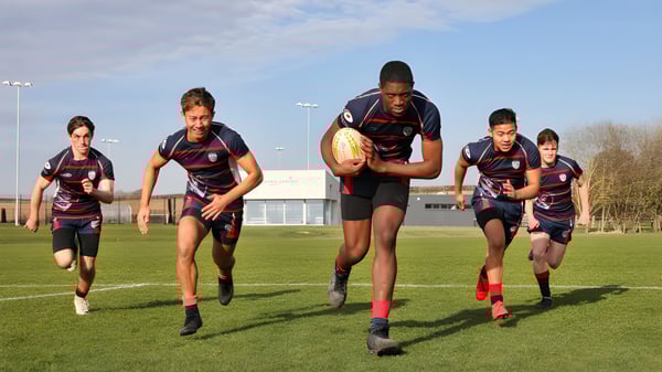 Alumnos de la Ashford School juegan rugby en un campo de césped frente a un edificio deportivo.