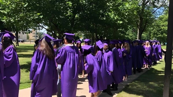 Un grupo de graduados en togas moradas y birretes camina por el campus verde de Assumption College Catholic High School.