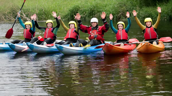 Un grupo de estudiantes del Athlone Community College rema con chalecos salvavidas coloridos en kayaks en un cuerpo de agua rodeado de vegetación verde.