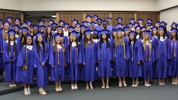 Un gran grupo de graduados de la Atlantic Shores Christian School está de pie en togas azules en una sala con paneles de madera.