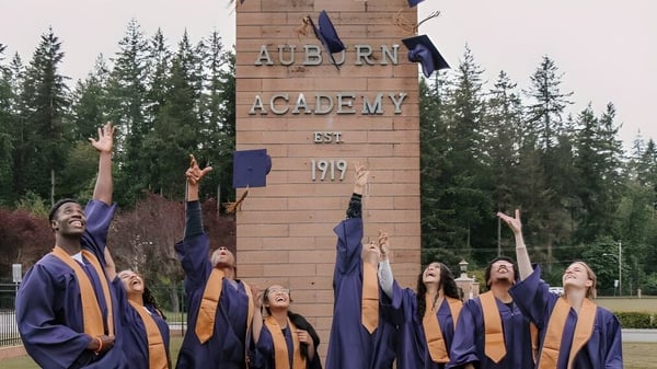 Un grupo de egresados de la Auburn Adventist Academy está de pie en túnicas violetas y naranjas frente al letrero de la escuela entre pinos.