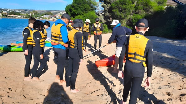 Un grupo de estudiantes del Avenues College está en trajes de neopreno en la playa cerca de un cuerpo de agua.