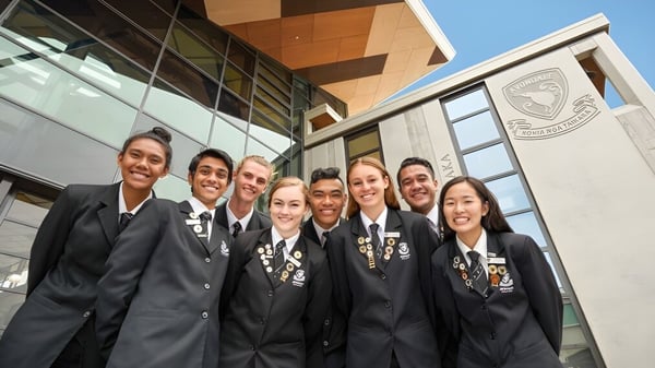 Un grupo de estudiantes en vestimenta académica formal está frente a un edificio con el escudo del Avondale College.