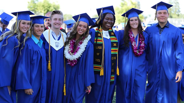 Un grupo de graduados en togas azules está afuera en el verde del campus de Bakersfield Christian.