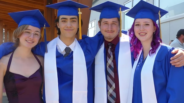 Cuatro egresados de la Ballenas Secondary School posan juntos en sus trajes de graduación en un entorno escolar.